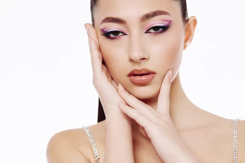 Beautiful woman with creative pink and purple eye makeup posing with hands on face. Studio portrait on white background. Concept of beauty fashion and artistic cosmetics.