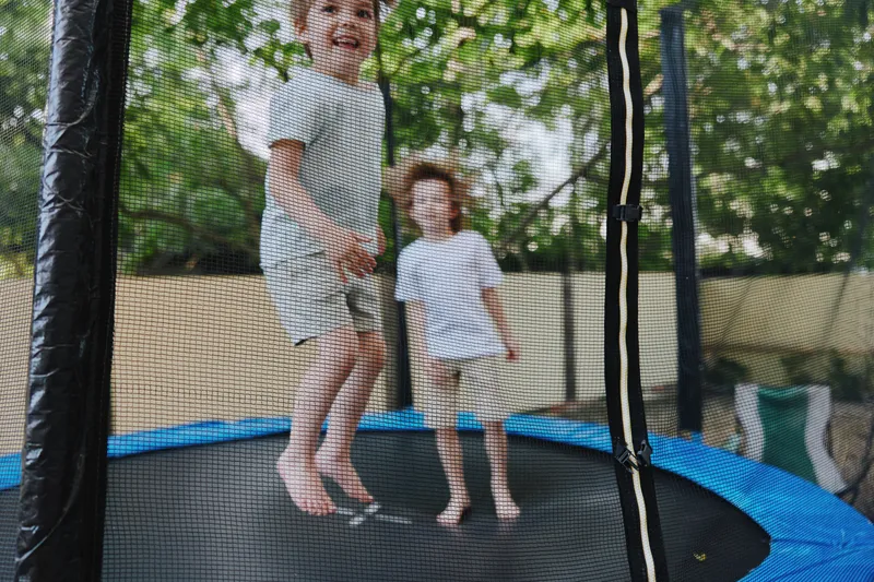 Two joyful boys bouncing on a trampoline, full of energy and laughter. The sunny day adds to the fun atmosphere of outdoor play, capturing childhood happiness.