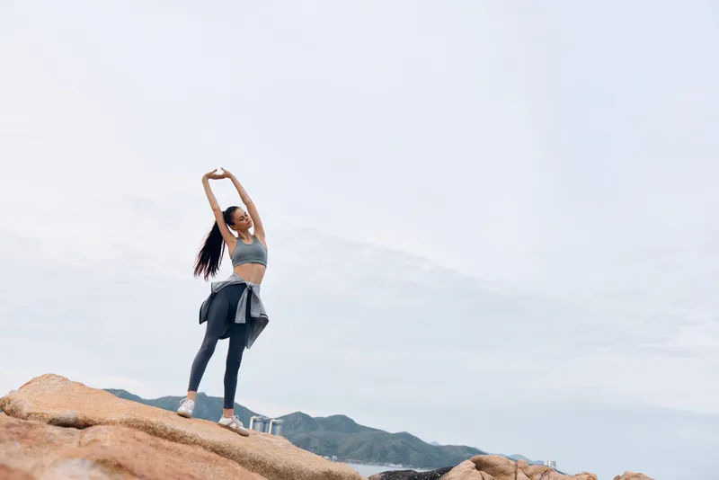 Young woman stretching on rocks by the sea, dressed in sportswear, enjoying nature and the outdoors in a tranquil setting, promoting healthy lifestyle and wellness