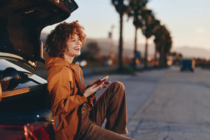 Woman sits by car holding smartphone on street at sunset, curly hair, relaxed lifestyle, urban roadside scene, warm evening light, casual portrait with open car door and traffic