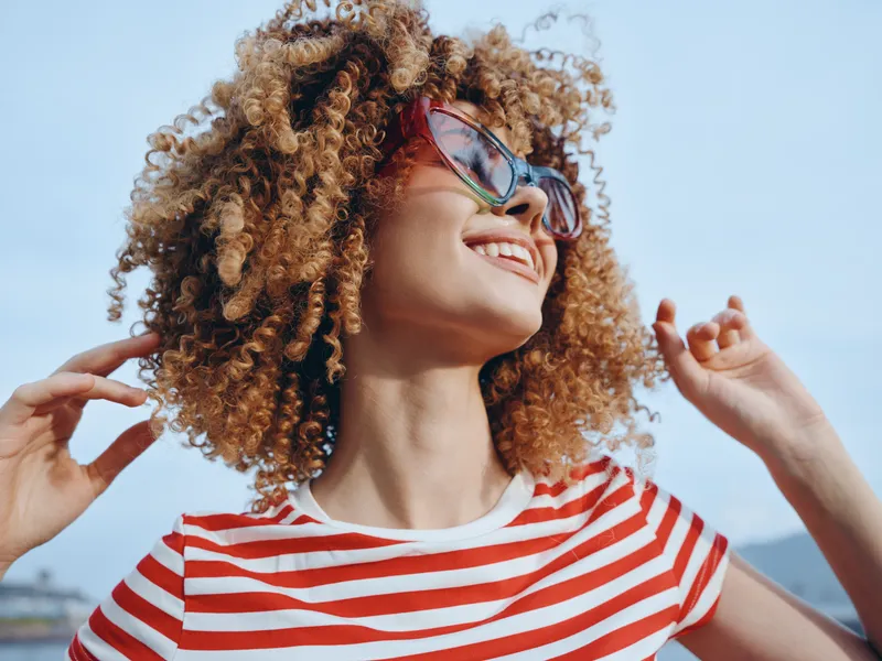 Young woman with curly hair smiles in sunglasses and a striped shirt outdoors, capturing carefree summer style and radiant confidence against a bright blue sky.