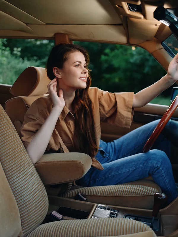 Young woman smiling while driving a vintage car, dressed casually in brown shirt and denim jeans, surrounded by a lush green background.