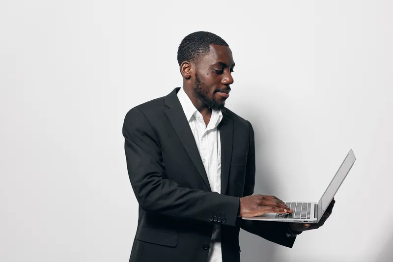 Black man with dark skin wearing black suit and white shirt working on laptop standing against white background. Business professional focused on technology and communication.