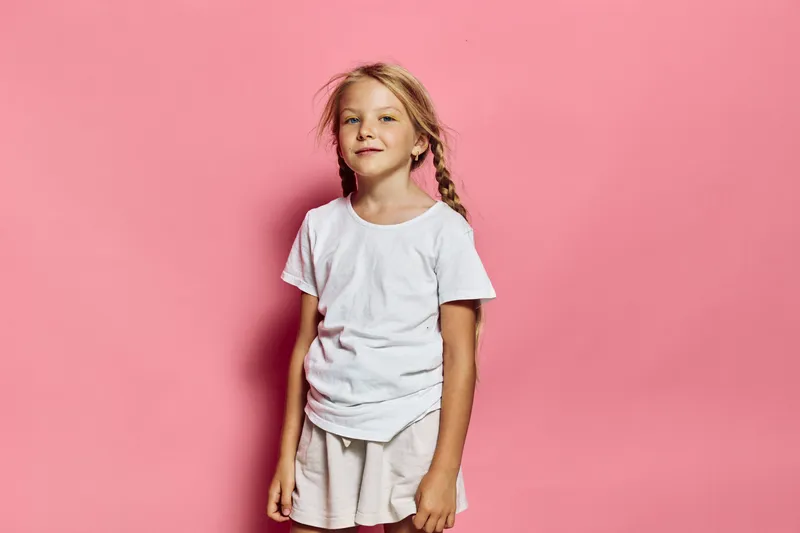 Young blonde girl with braided hair wearing a white t-shirt and beige shorts stands confidently against a pink background. Studio shot with casual summer outfit concept.