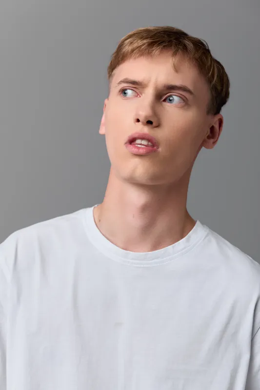 Man with a puzzled expression looks away, wearing a casual white shirt in a simple studio setting, conveying thoughtfulness, uncertainty, and curiosity during a moment of decision and reflection