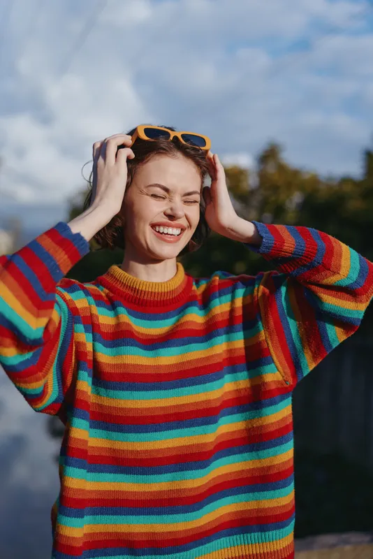 Young woman in colorful striped sweater smiling with sunglasses on her head outdoors, casual portrait of a joyful twenties model laughing and enjoying warm natural light.
