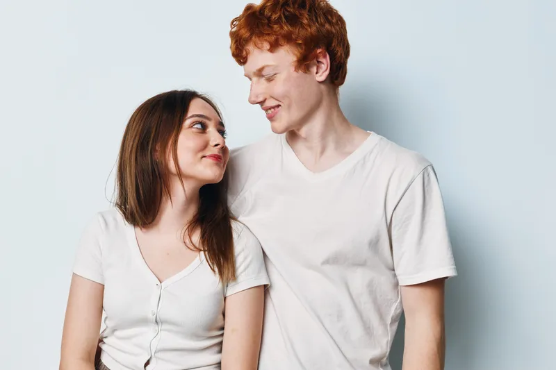 Young couple standing against light background looking at each other smiling casual clothing together. Man and woman with red hair and brown hair posing and showing affection.
