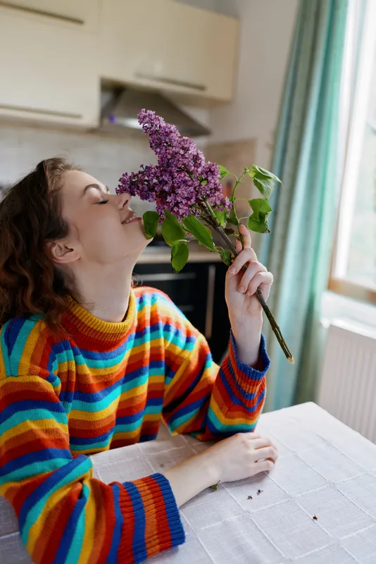Young woman in a colorful striped sweater smells a lilac bouquet at a kitchen table, capturing a warm intimate home moment.