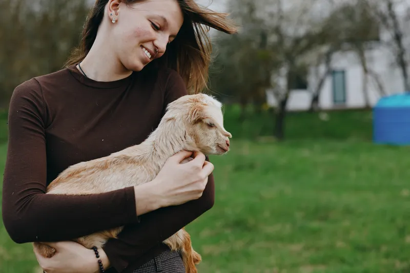Happy woman outdoors holds a gentle goat kid, smiling warmly, wearing a brown sweater, in a green park with trees, creating a wholesome lifestyle moment focused on care, affection, and bonding with a