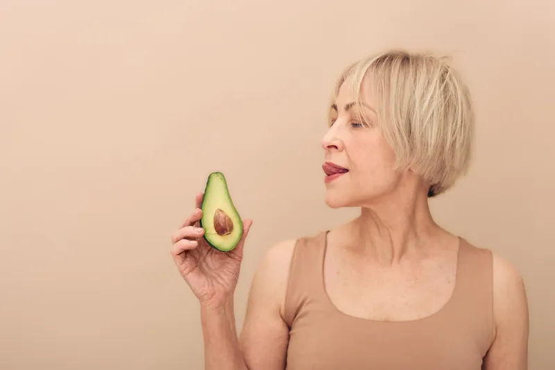 Blonde mature woman with short hair holds half of fresh avocado near face, standing against beige background. Concept of healthy eating, natural beauty, wellness, and skin care routine.