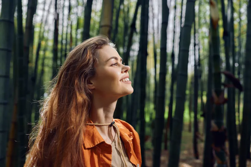 Woman smile in bamboo forest with nature sunlight on her face, profile portrait of a redhead in an orange jacket enjoying peaceful outdoors, relaxation and joyful serenity.