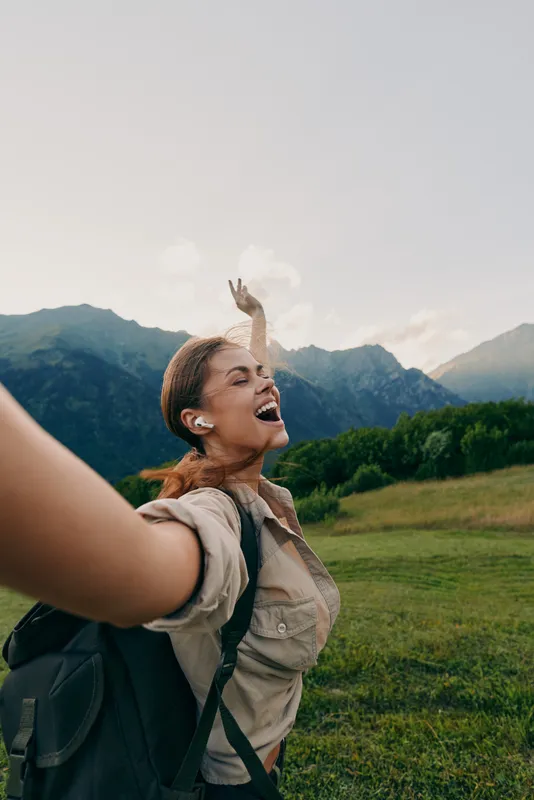 Joyful woman hiking outdoors with backpack, arms raised in celebration, amid green fields and mountains, conveying adventure, freedom, and vibrant outdoor lifestyle.