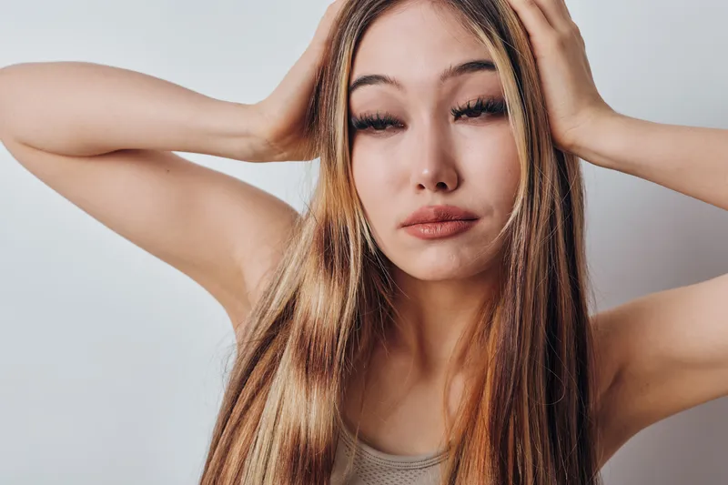 Woman with long hair holding head with hands and closed eyes showing stress or headache in studio with plain background. Female person expressing tiredness or discomfort in closeup portrait.
