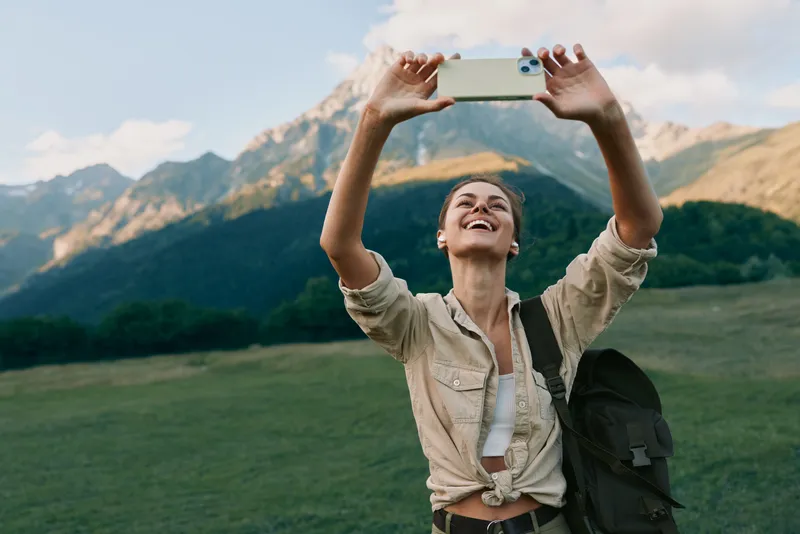 Young woman with a backpack takes a selfie with a smartphone in a mountain meadow under a partly cloudy sky, expressing joy and freedom during an outdoor adventure.