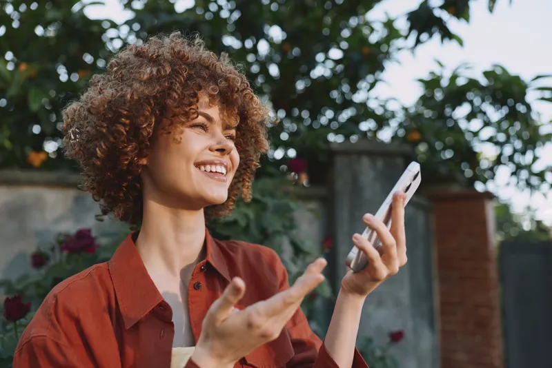 A young woman with curly hair smiles while checking her smartphone outdoors in a sunlit garden, wearing a rust colored shirt and enjoying a relaxed moment