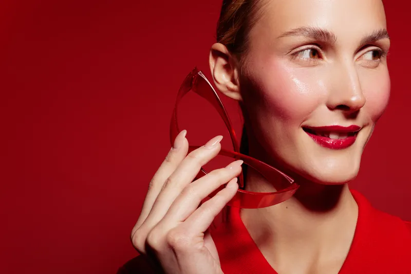 Close-up of a smiling woman with red lipstick holding a red heart-shaped object near her face on a red background. Concept of love, beauty, and romantic emotions.