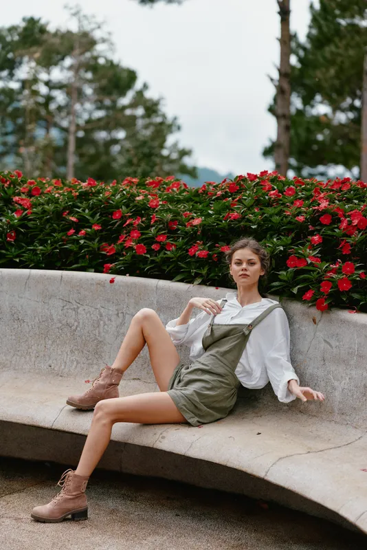 Young female model in stylish overalls, sitting gracefully against lush flowers, capturing a relaxed spring vibe with a hint of fashion uniqueness