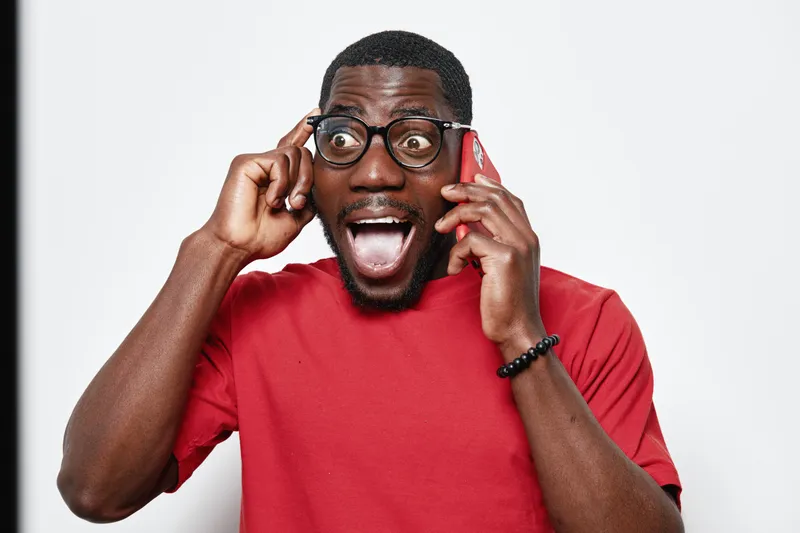 African American man with dark skin wearing glasses and red shirt talking on smartphone with excited expression and open mouth on white background. Communication, technology and emotion.