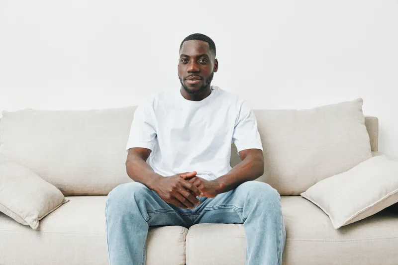 Confident African American man sitting on a light colored couch, wearing a simple white t shirt and jeans, reflecting casual style and relaxation