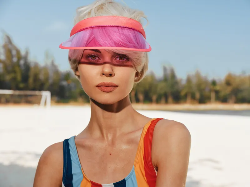 Vibrant model outdoors wearing a color block swimsuit and translucent visor, sunlit skin and bold fashion statement, close portrait highlighting style, summer mood, playful confidence, beach setting.