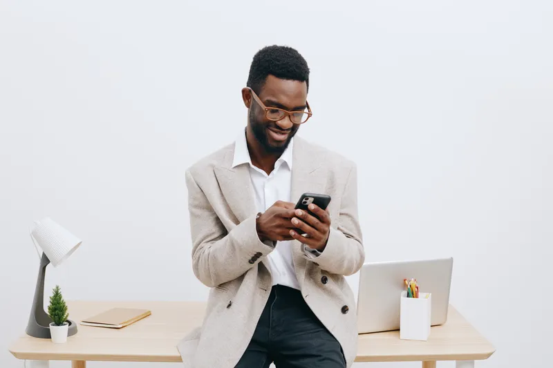 Smiling young African American man in casual blazer and glasses using smartphone at modern office workspace with laptop, pen holder, and decorative plants, indoor business casual lifestyle concept, fr
