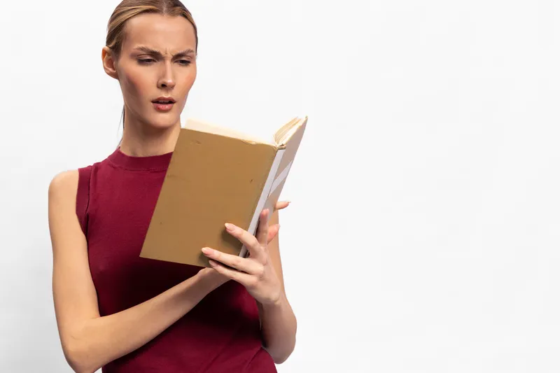 Confused woman in red sleeveless top reads a book with puzzled expression isolated on white background. Concept of misunderstanding, concentration, and learning challenges.