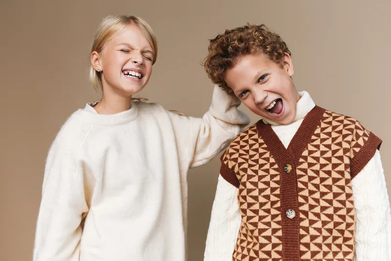 Two joyful children laugh together, wearing soft knit sweaters against a warm neutral backdrop, capturing a moment of carefree friendship, playfulness, and lighthearted warmth.