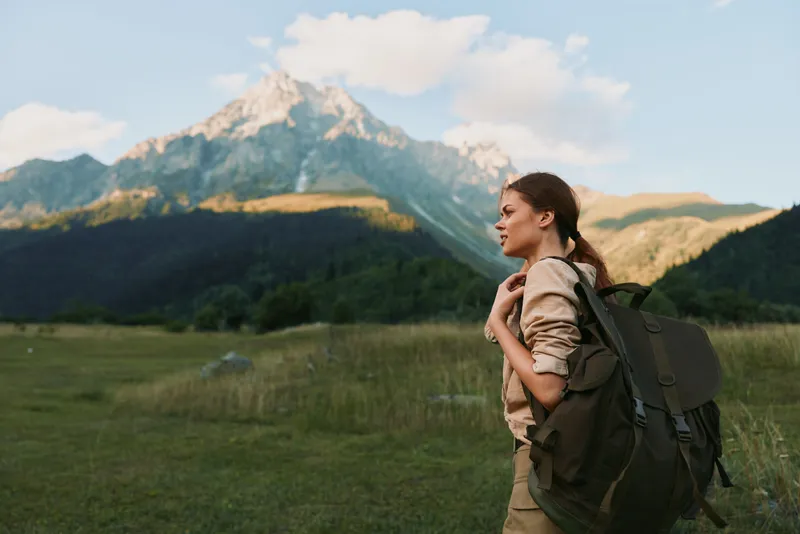 Young adult woman hiking in a mountain landscape with a large backpack, enjoying outdoor adventure and a clear sky.