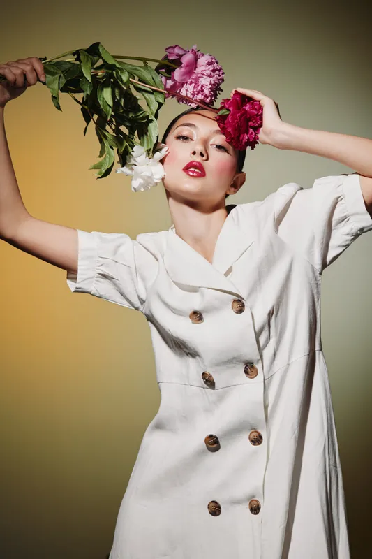 Fashionable woman in white dress holds pink and white peonies near her head with confident expression. Beauty concept with creative flowery composition on gradient background.