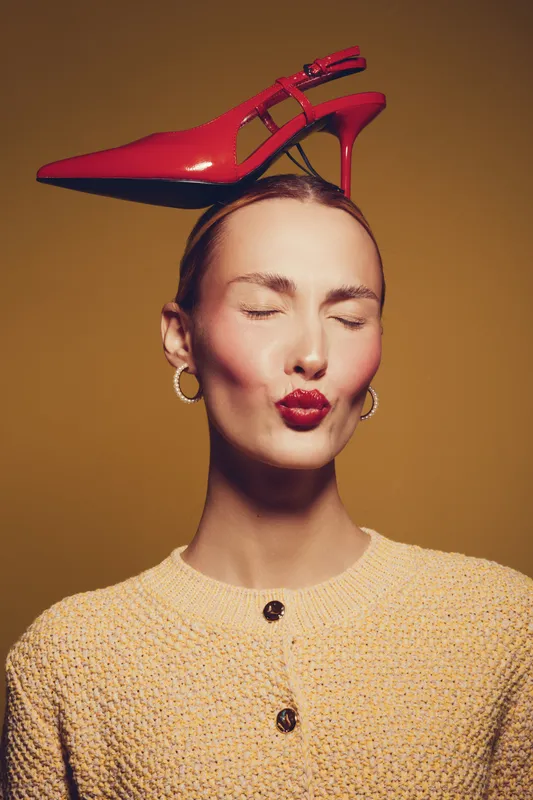 Young woman with closed eyes and red lipstick balances red high heel shoe on her head. She wears a yellow knitted sweater and gold hoop earrings on brown background. Fashion concept.