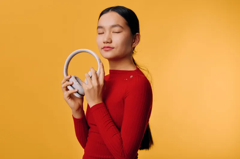 Young woman in red sweater enjoying music with closed eyes holding wireless headphones near face against yellow background. Relaxation, leisure and sound experience with modern audio device.