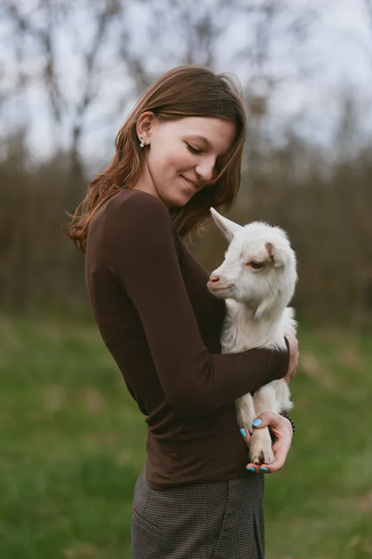 Cheerful woman outdoors, embracing a gentle goat kid, warm sunlight, soft breeze, relaxed posture, intimate connection, natural colors, countryside backdrop