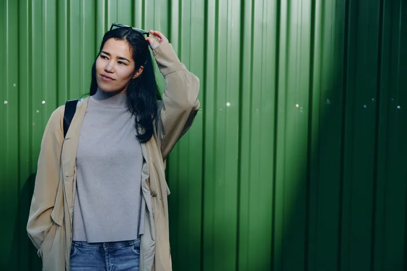 Woman standing near a green wall, wearing a beige coat and gray turtleneck, adjusting hair, casual urban style, accessorized with a backpack, relaxed pose and soft natural lighting.