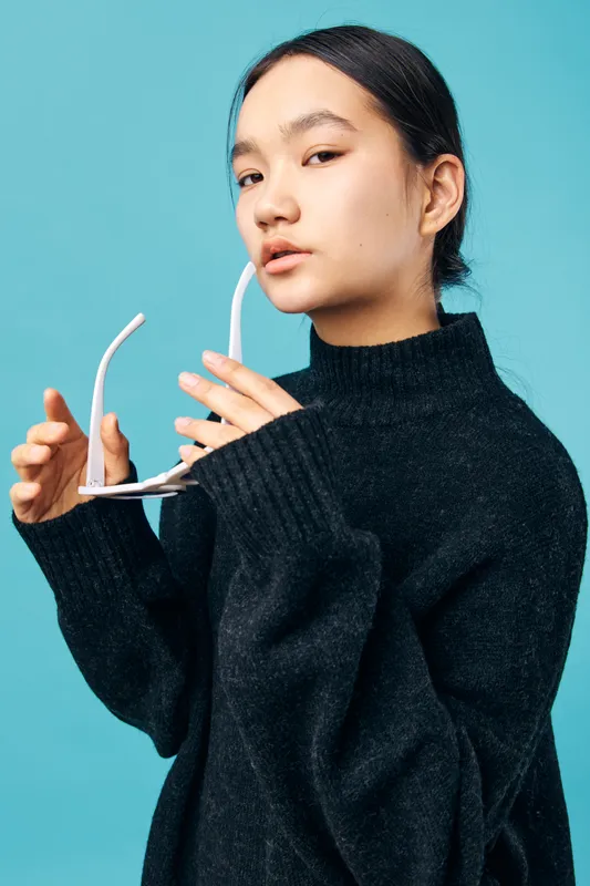 Young woman with black sweater holding white eyeglasses, posing on blue background with serious expression, fashion portrait of female beauty and casual style with minimal accessories.