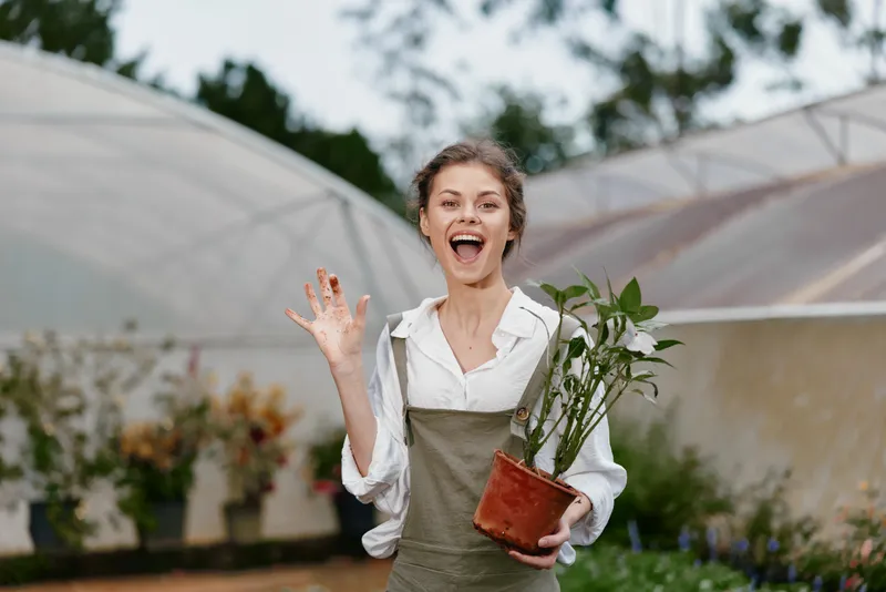 Joyful young woman holding a potted plant in a greenhouse, showcasing enthusiasm for gardening and a healthy lifestyle, surrounded by greenery and vibrant colors