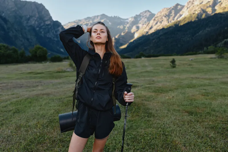 A confident hiker stands in a scenic mountain meadow with a backpack and trekking pole, embracing the outdoors and fresh alpine air under clear blue skies.