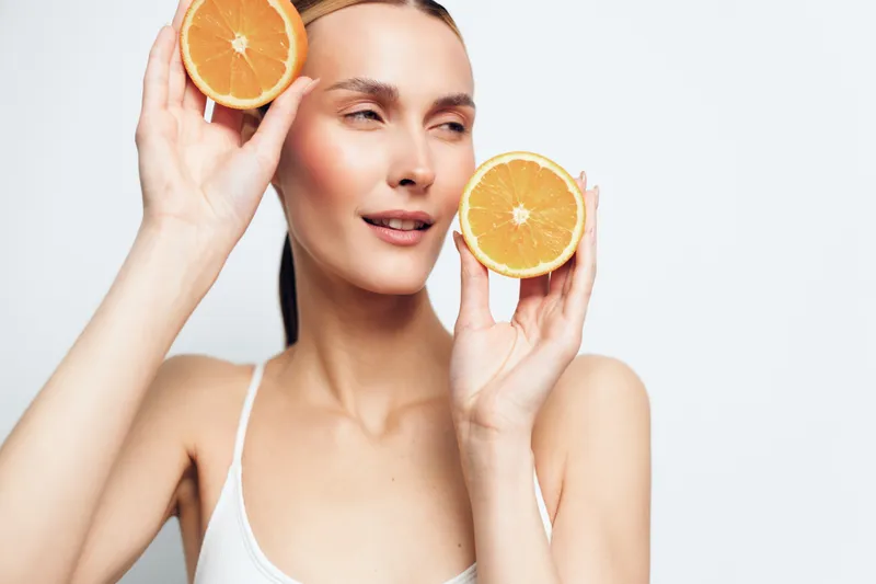Young woman with clear skin holds two halves of a fresh orange near her face while smiling softly against a white background, conveying freshness and natural beauty.