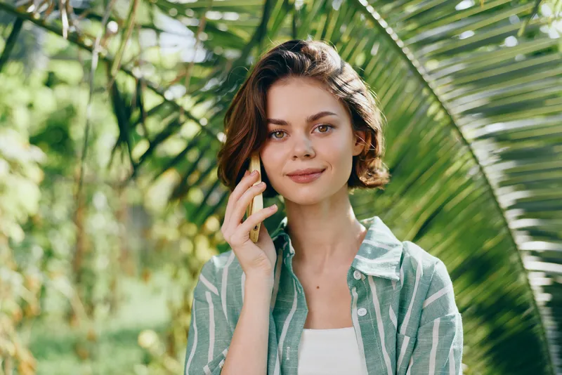 Portrait of a young woman outdoors among tropical greenery, with a soft smile and casual striped shirt under natural light.