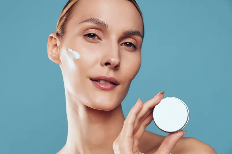 A young woman with light skin applies cream to her face in a close up studio shot, highlighting a skincare routine and quiet self care moment.