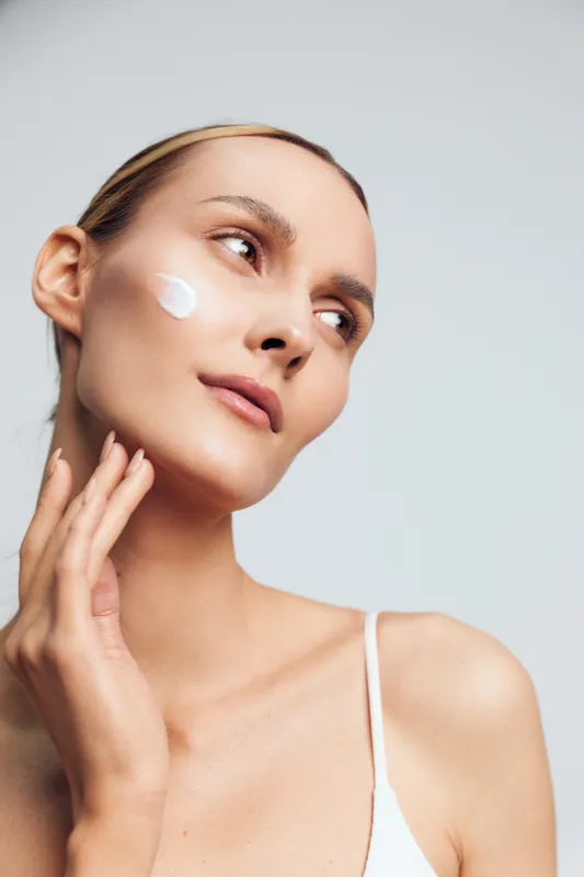 Woman applying face cream skincare self care close up studio lighting, relaxed expression with white moisturizer on cheek, soft studio portrait on light background, healthy skin wellness routine