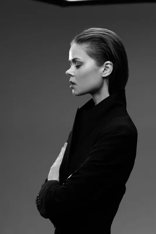 Black and white profile portrait of a woman in a black coat with folded arms and a serious expression