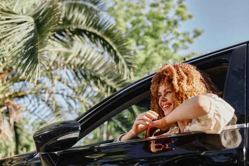 smiling woman with curly hair leaning out of black car window outdoors, tropical background, sunny day, casual clothing, positive mood