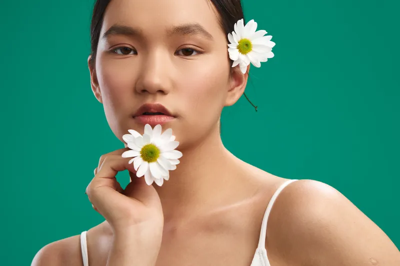 asian woman gazes softly at the camera, delicate daisies tucked in her hair and near her chin, creating a fresh beauty portrait with a calm, studio vibe.
