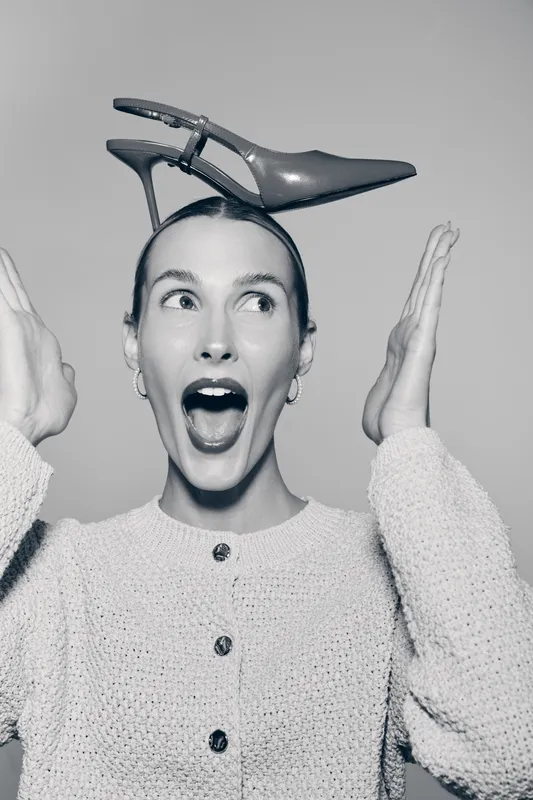 Happy Caucasian woman with fair skin holds high heel shoe on head, surprised expression, wearing knitted sweater. Concept of fashion, fun, and creativity in studio portrait.