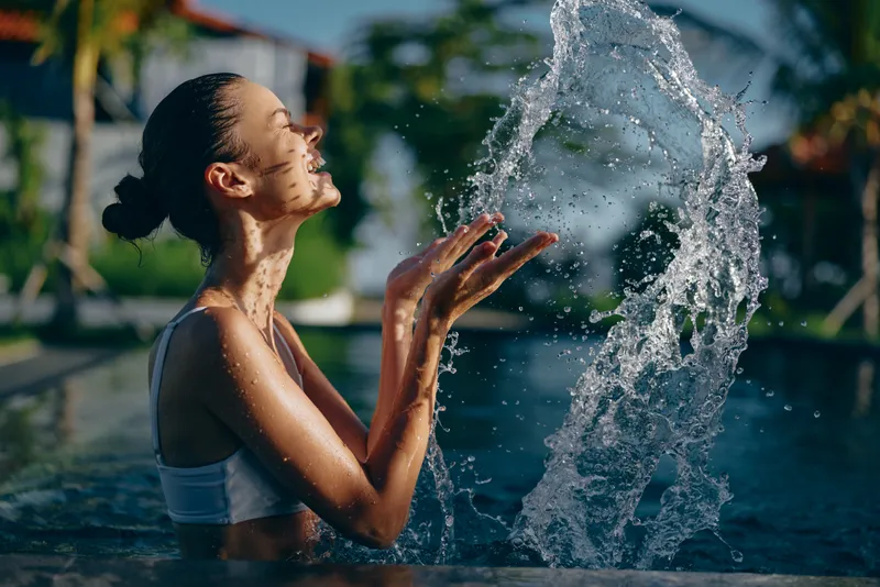 A woman in a swimsuit plays with water in a pool, capturing a candid lifestyle moment of refreshment, fitness, and carefree outdoor relaxation.