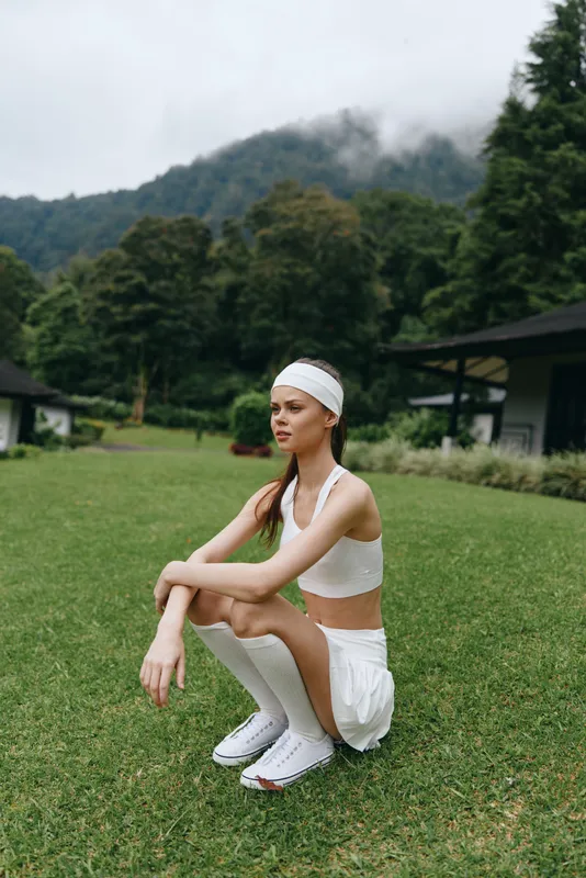 Young woman in white athletic wear squatting on grass, showcasing fitness lifestyle in a tranquil outdoor setting surrounded by greenery and mountains