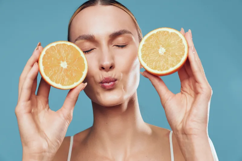 Young woman with closed eyes holding two orange halves near face, puckering lips, fresh citrus fruit, smooth skin, blue background, natural beauty, healthy lifestyle concept