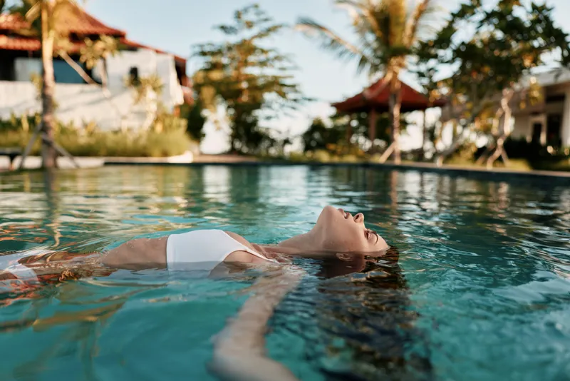 Relaxed woman floating on back in outdoor pool, eyes closed, wearing white swimsuit, tropical resort setting, calm water, sunny day, peaceful moment