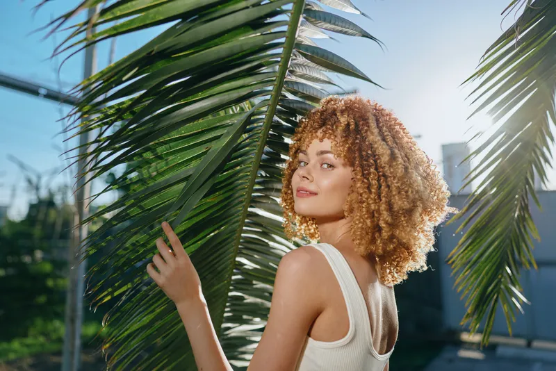 curly woman in white tank top standing near palm leaves under bright sunlight outdoors, natural light, summer mood, casual style, warm tones