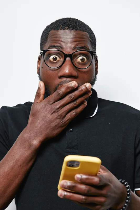 Black man with dark skin and glasses looking shocked while holding yellow smartphone in hand. Surprised African American male with beard wearing black polo shirt on white background.